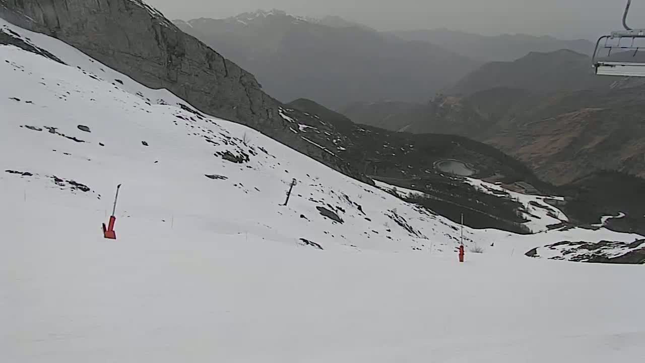 A snow-covered ski slope descends into a valley on an overcast day, with two orange ski markers visible.