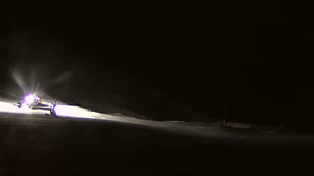At night, under a dark sky, snow groomers illuminate a snow-covered slope with bright lights.