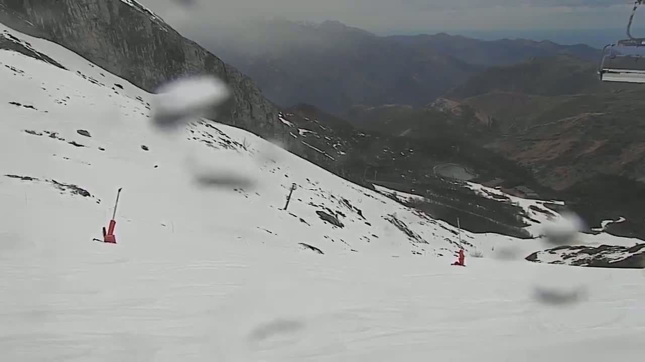 A snowy mountain slope with two skiers visible in the distance, viewed through rain-covered glass on a cloudy day.