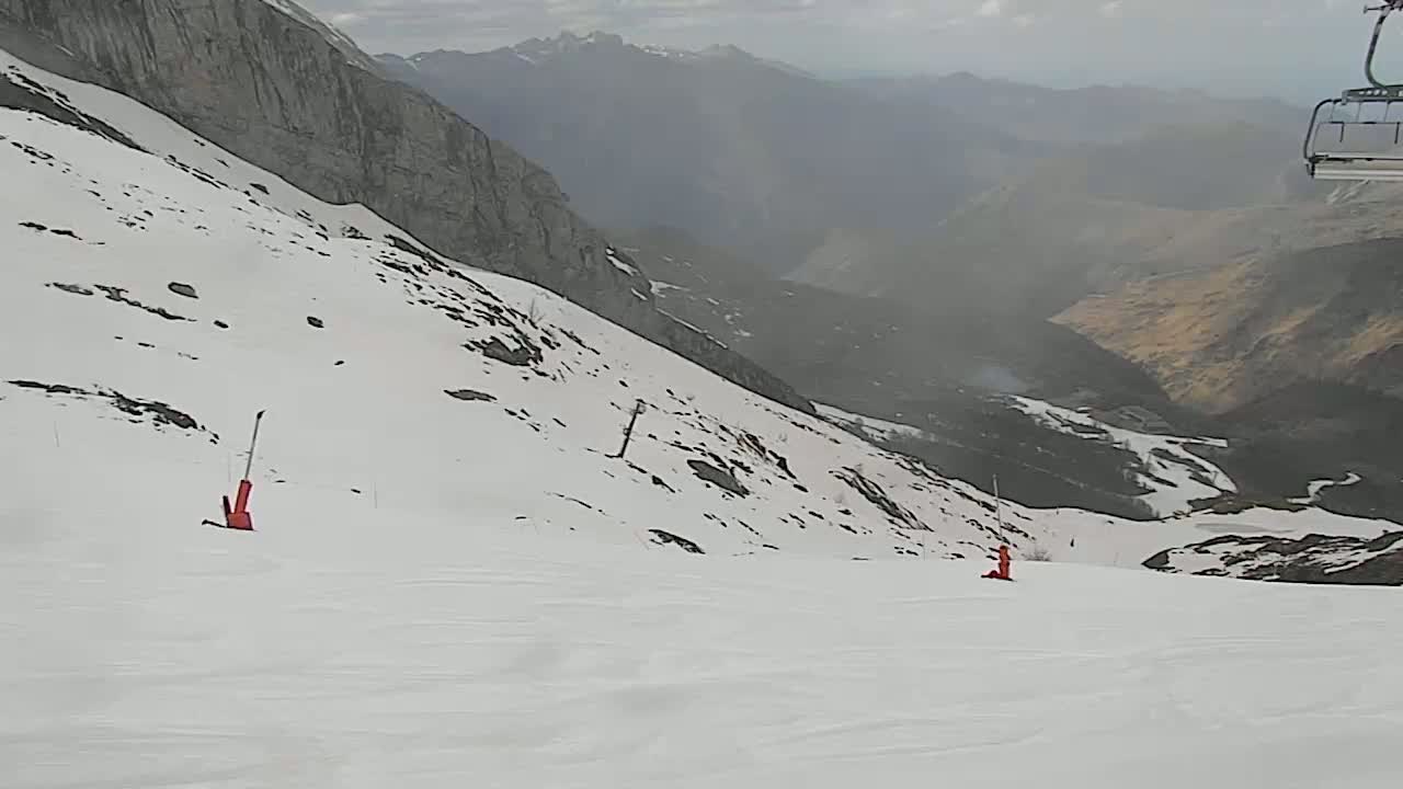 A snow-covered mountain slope with two skiers visible, overlooking a valley in hazy weather conditions.