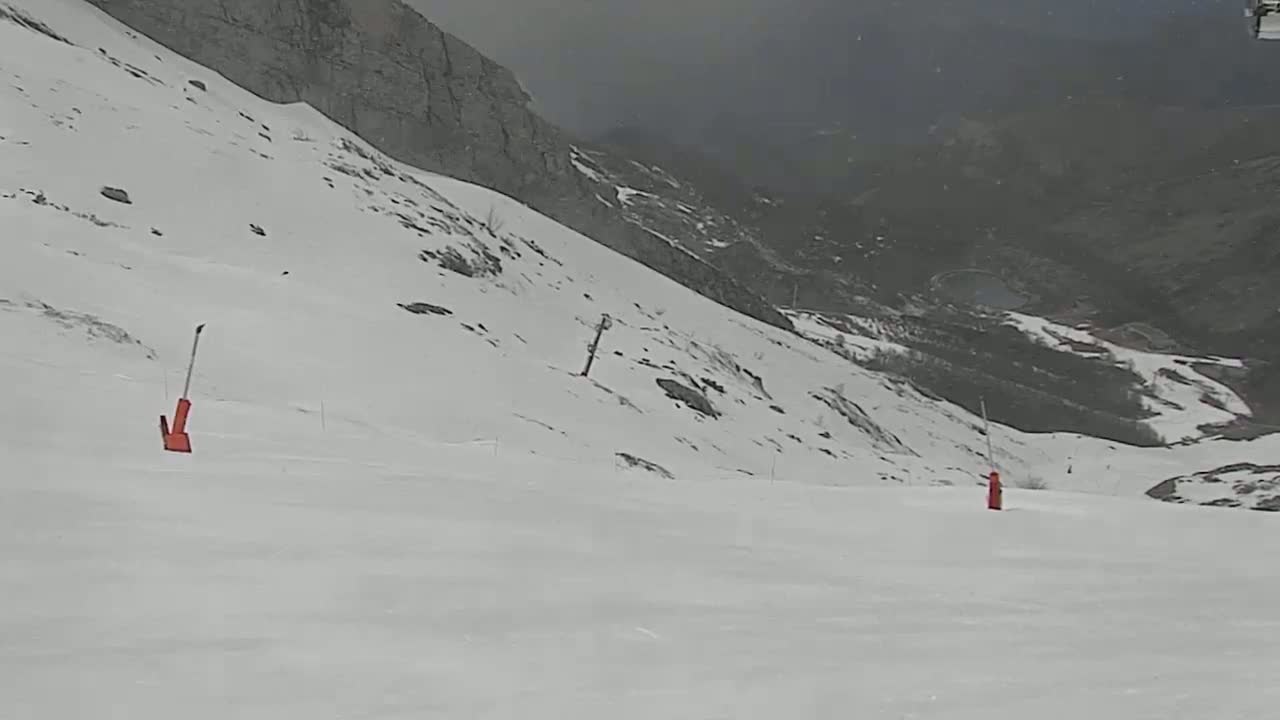 A snow-covered mountain slope with two orange ski markers is visible under an overcast sky.
