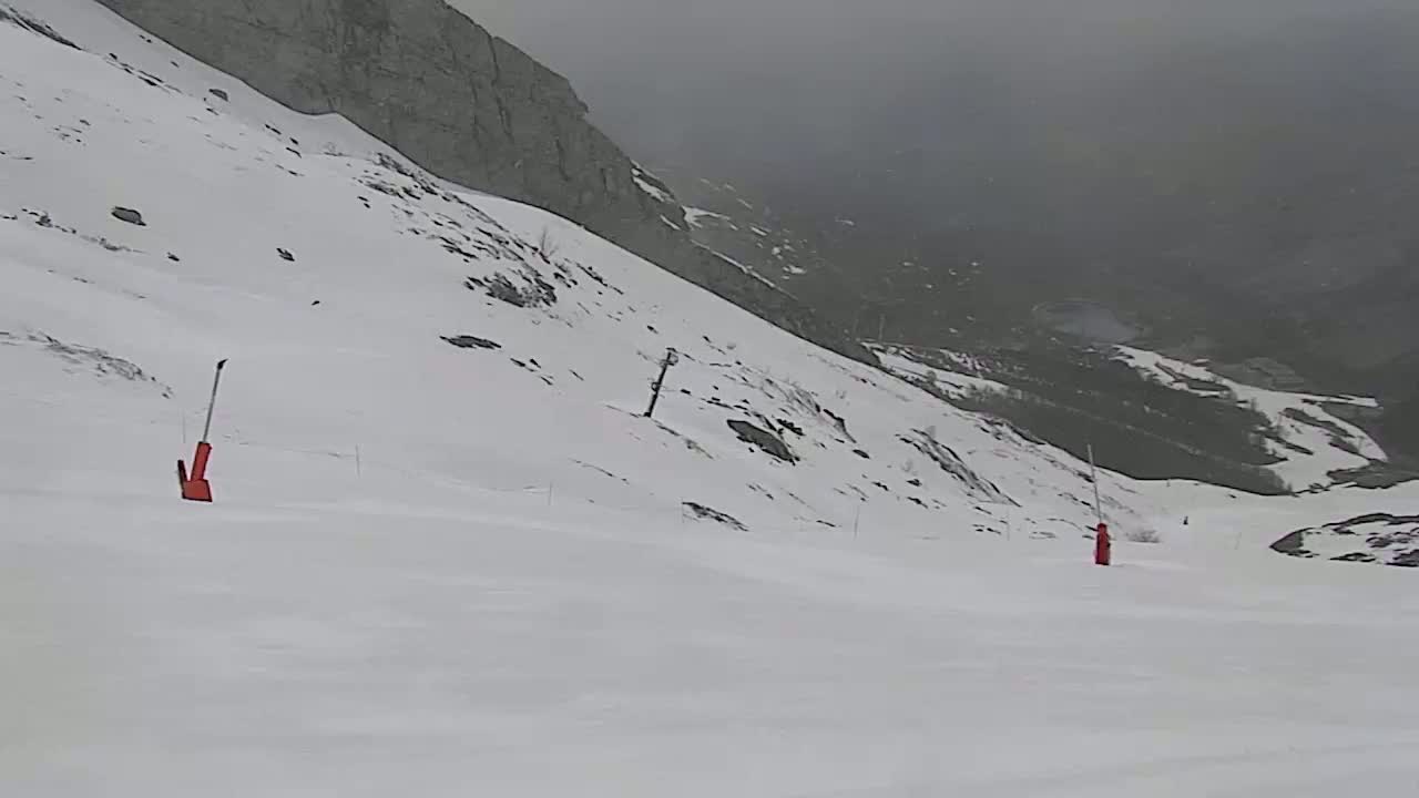 A snow-covered ski slope descends into a foggy valley, with two orange markers visible on the piste.