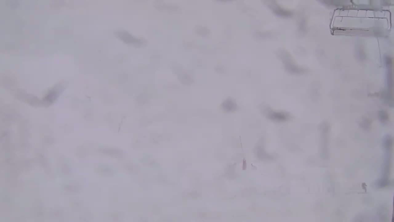 A snowy, blurry scene shows a portion of a ski lift and faint figures in the heavy snow.