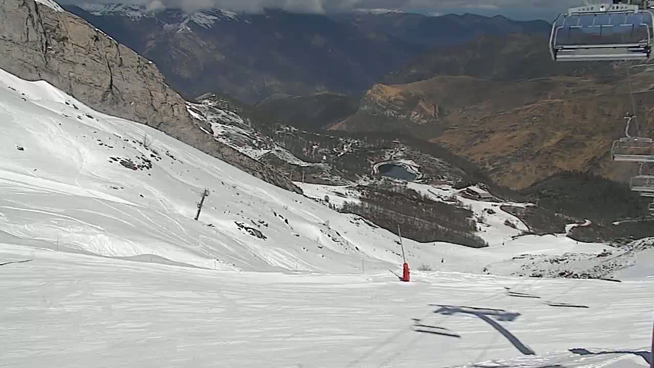 A snowy ski slope descends toward a valley with a small lake, under partly cloudy skies.