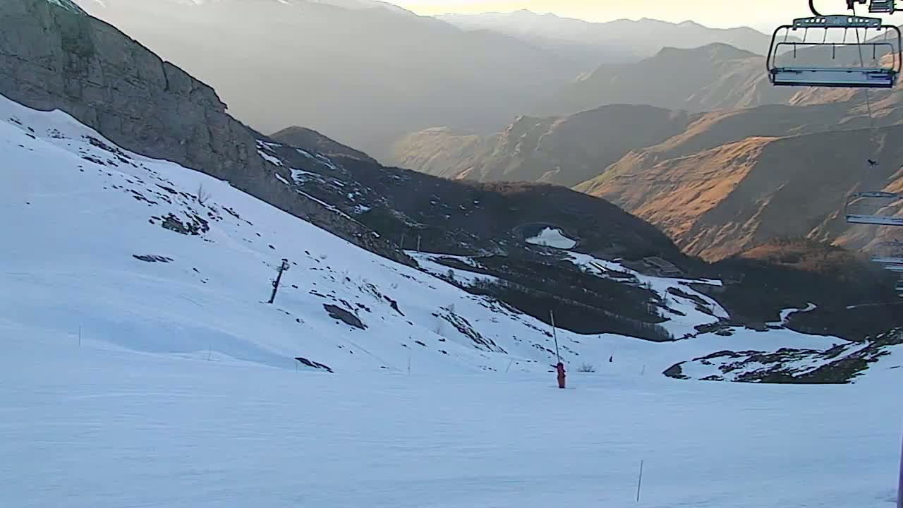 A snowy mountain slope with a ski lift and a skier visible in the distance under a hazy sky.