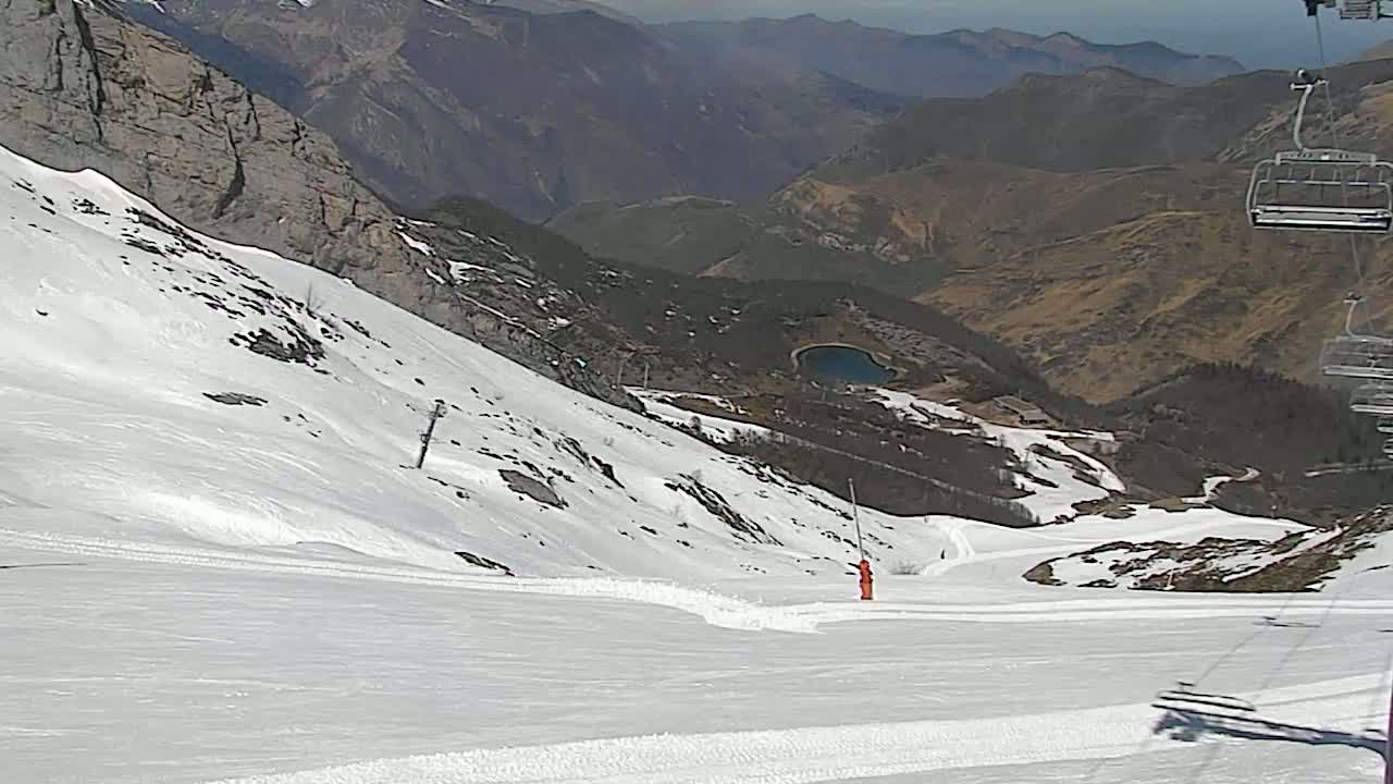 A snowy mountain slope with a person in orange standing near a ski lift, overlooking a valley with a small lake under a partly sunny sky.