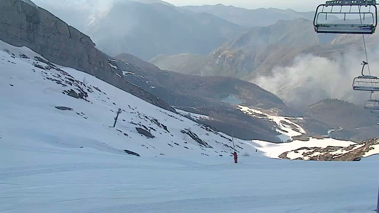 A skier stands on a snow-covered mountain slope overlooking a valley with some smoke or haze in the distance, under a mostly clear sky.