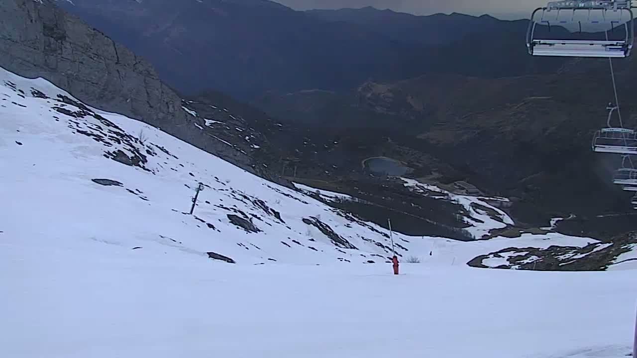 A skier is seen on a snowy mountain slope overlooking a valley, with a chairlift in the upper right corner.