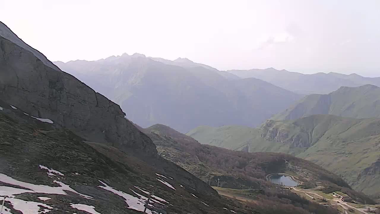 A mountain range stretches into the hazy distance, with patches of snow visible on a closer, rocky slope and a small lake nestled in a valley below.