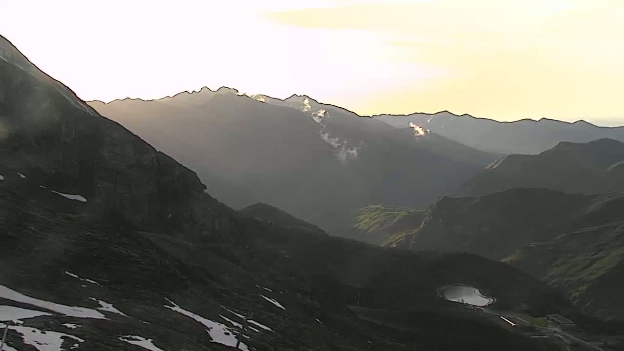 A mountain range at sunset, with patches of snow visible on the lower slopes, under a mostly clear sky.