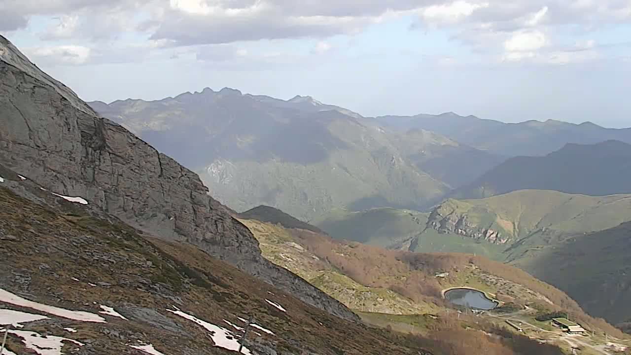 A mountainous landscape with patches of snow, a small lake, and partly cloudy skies is seen from a high vantage point.
