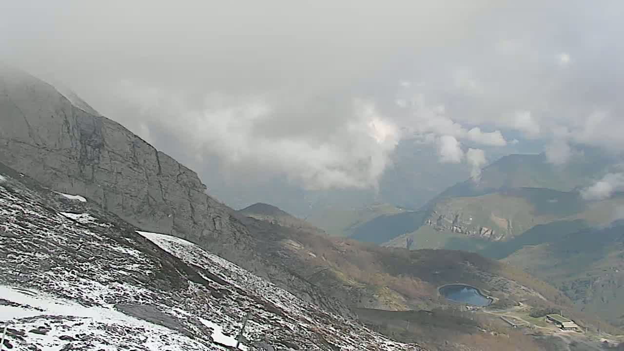 A snow-dusted mountainside overlooks a valley shrouded in clouds, with a small lake visible in the distance.