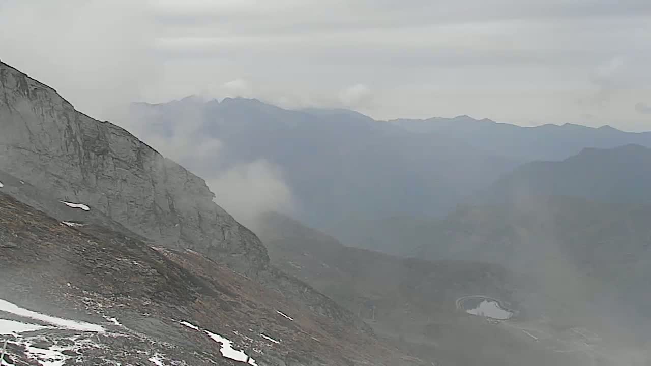 A hazy, overcast day reveals a steep, rocky mountainside sloping down to a distant valley of rolling hills and a small lake, partially obscured by fog.