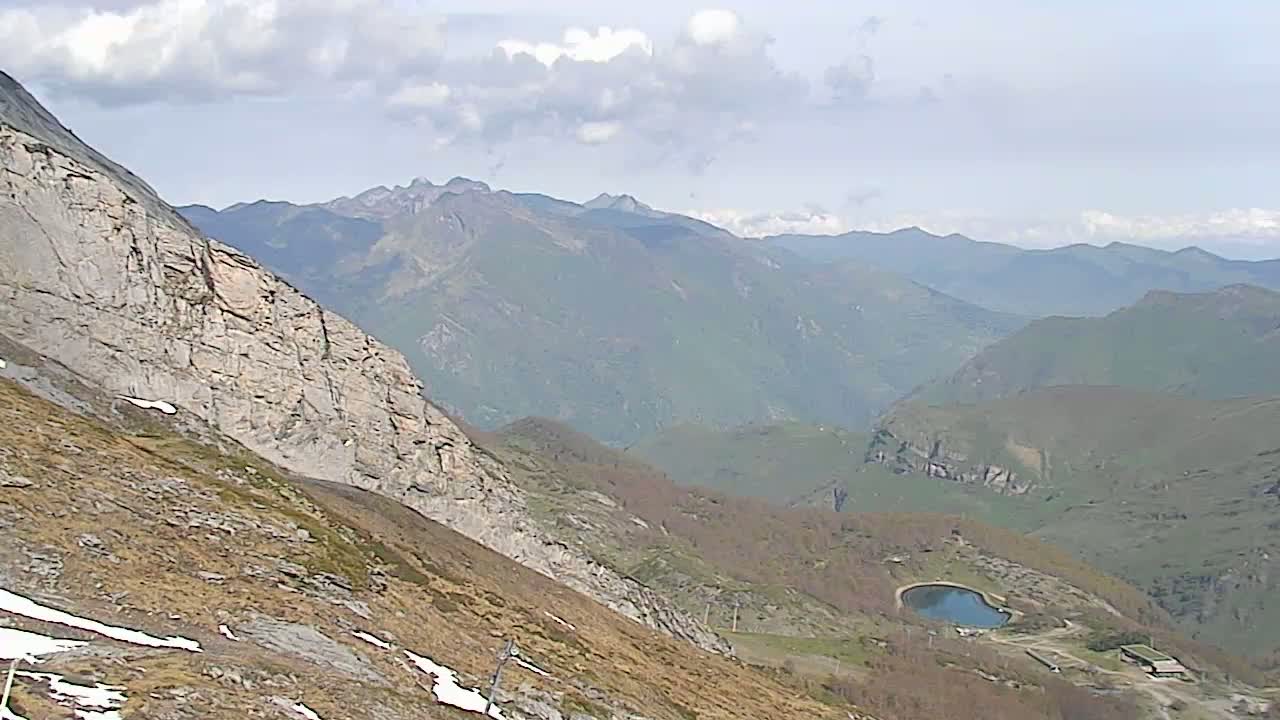 A mountainous landscape under a partly cloudy sky shows a steep, rocky slope in the foreground with patches of snow and brown vegetation, overlooking a valley with a small reservoir and distant, hazy peaks.