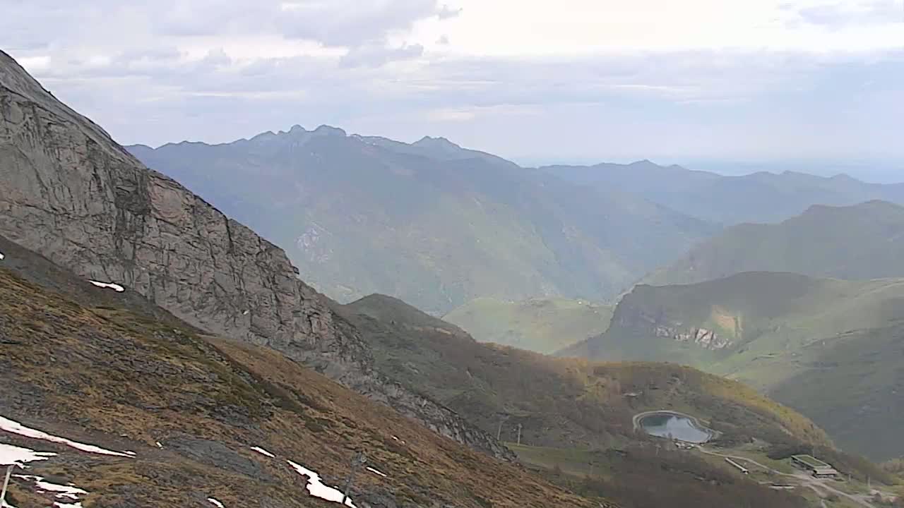 A mountainous landscape under a cloudy sky shows a steep, rocky cliff face in the foreground, leading down to a valley with a small reservoir and distant hazy peaks.