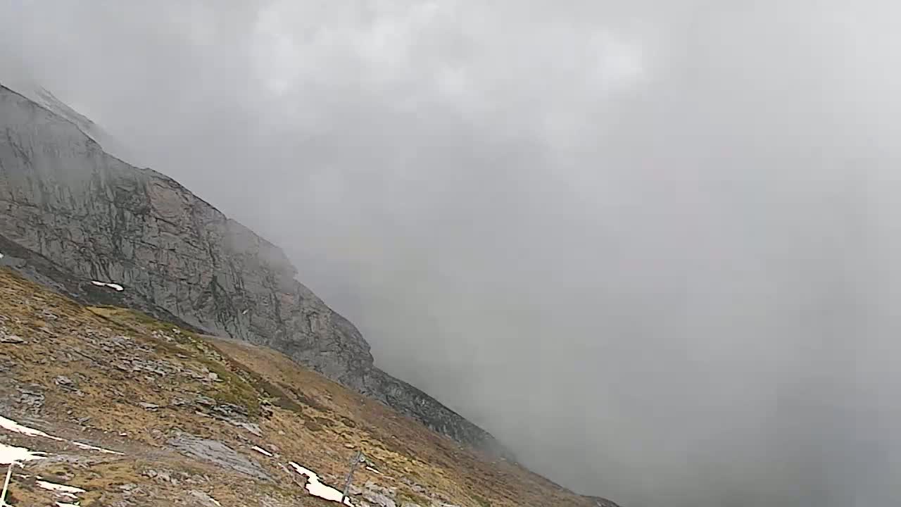 A rocky mountainside, partially covered in patchy vegetation and snow, is shrouded in thick fog.