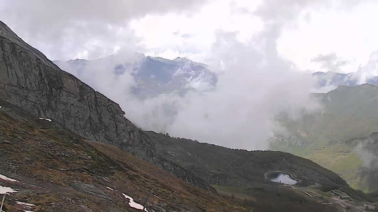 A mountainous landscape, partially obscured by fog, shows a rocky cliff face on the left and a valley below with a small reservoir.