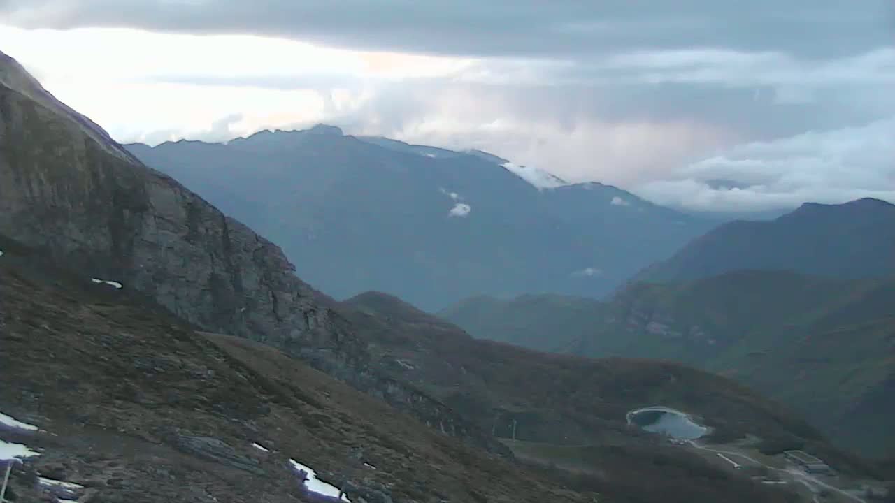 A view from a mountaintop shows a cloudy sky over a series of distant, hazy mountains, with a small lake visible in a valley below.