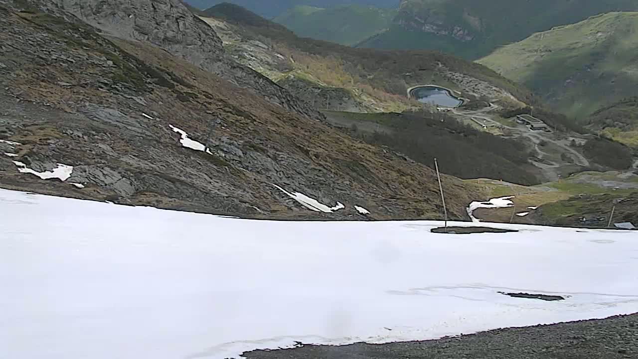 A mostly snow-covered mountain slope overlooks a valley with a small lake and some structures, under overcast skies.