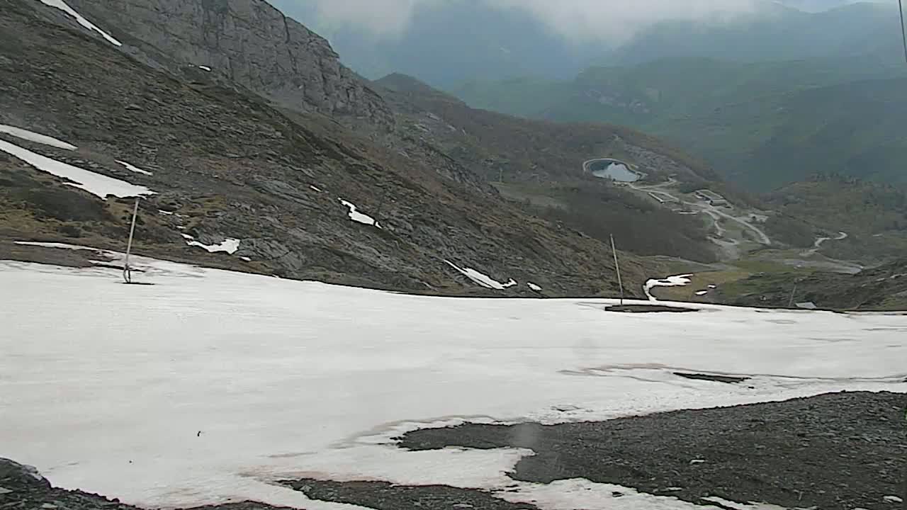 A mostly snow-covered mountainside slopes down to a valley with winding roads and a small body of water under an overcast sky.
