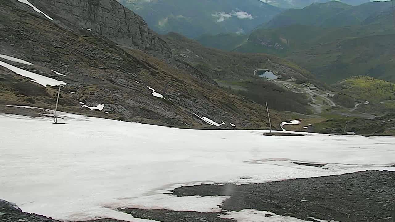 A snow-covered mountain slope descends toward a valley with a small lake and winding roads, under an overcast sky.
