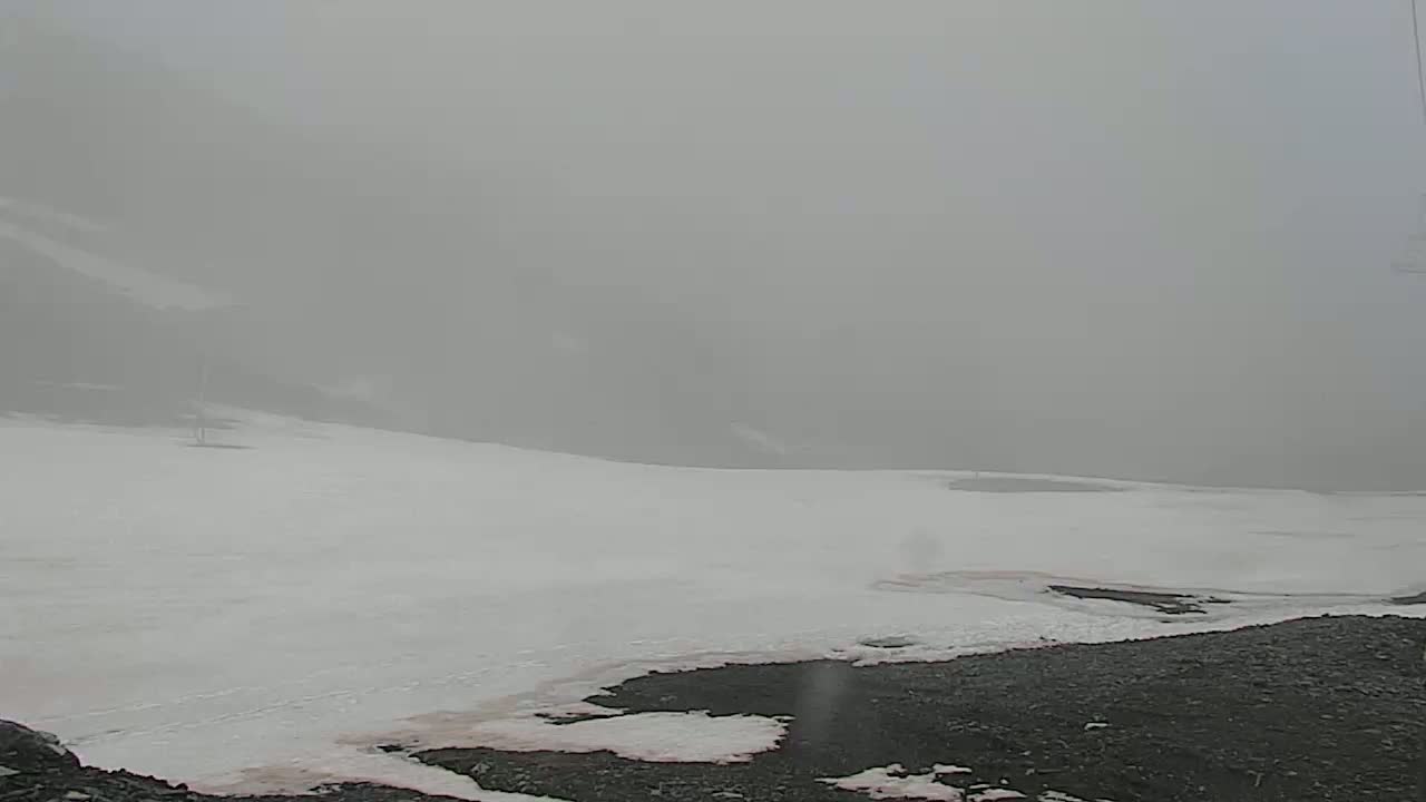 A snowy, foggy landscape shows a mostly flat expanse of snow with dark patches of ground visible at the bottom,  under a heavy overcast.