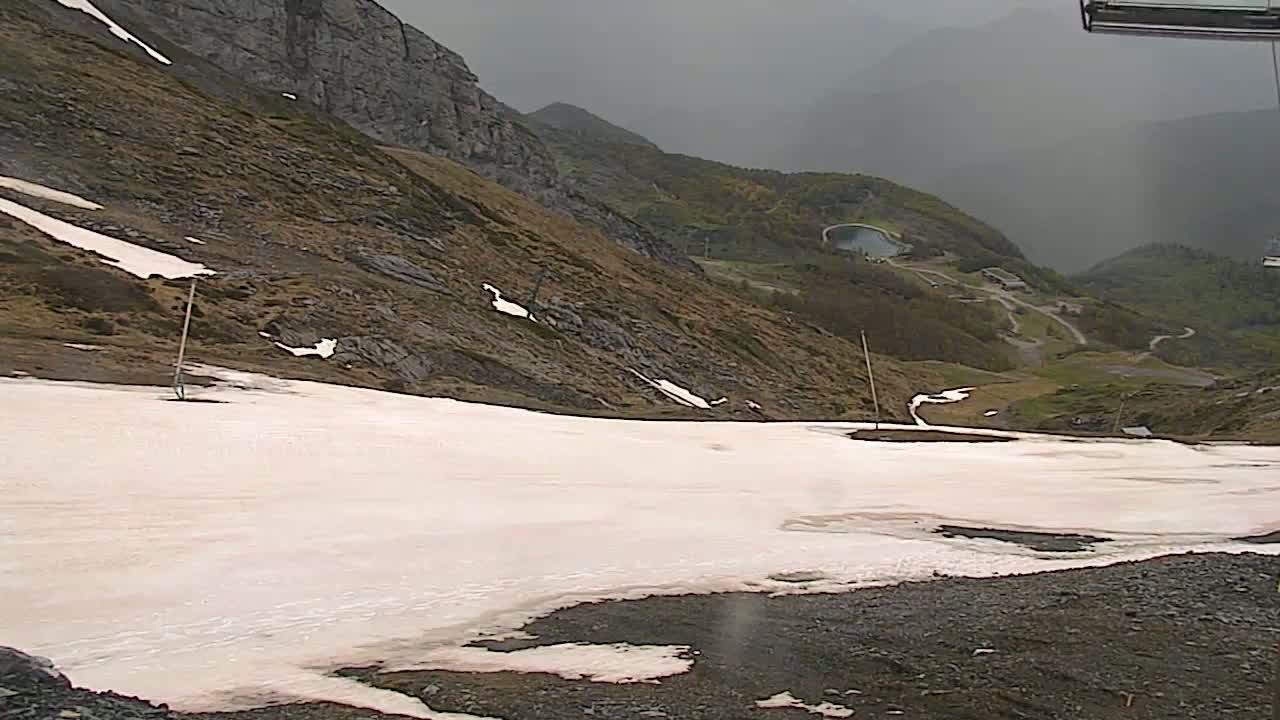 A large expanse of melting snow sits atop a dark-colored mountain slope overlooking a valley with some green vegetation and a small body of water under an overcast sky.