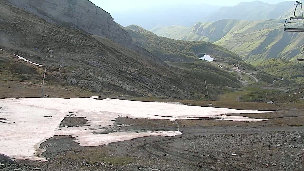 A rocky mountainside with patches of melting snow overlooks a valley with a ski resort in the distance under a clear sky.