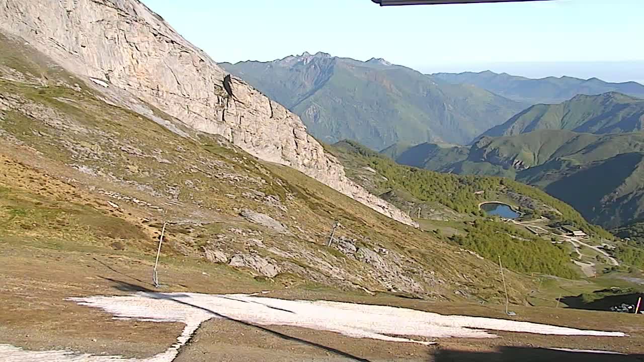A mountain slope with patches of snow, overlooking a valley with a small lake and distant mountains under a clear sky.