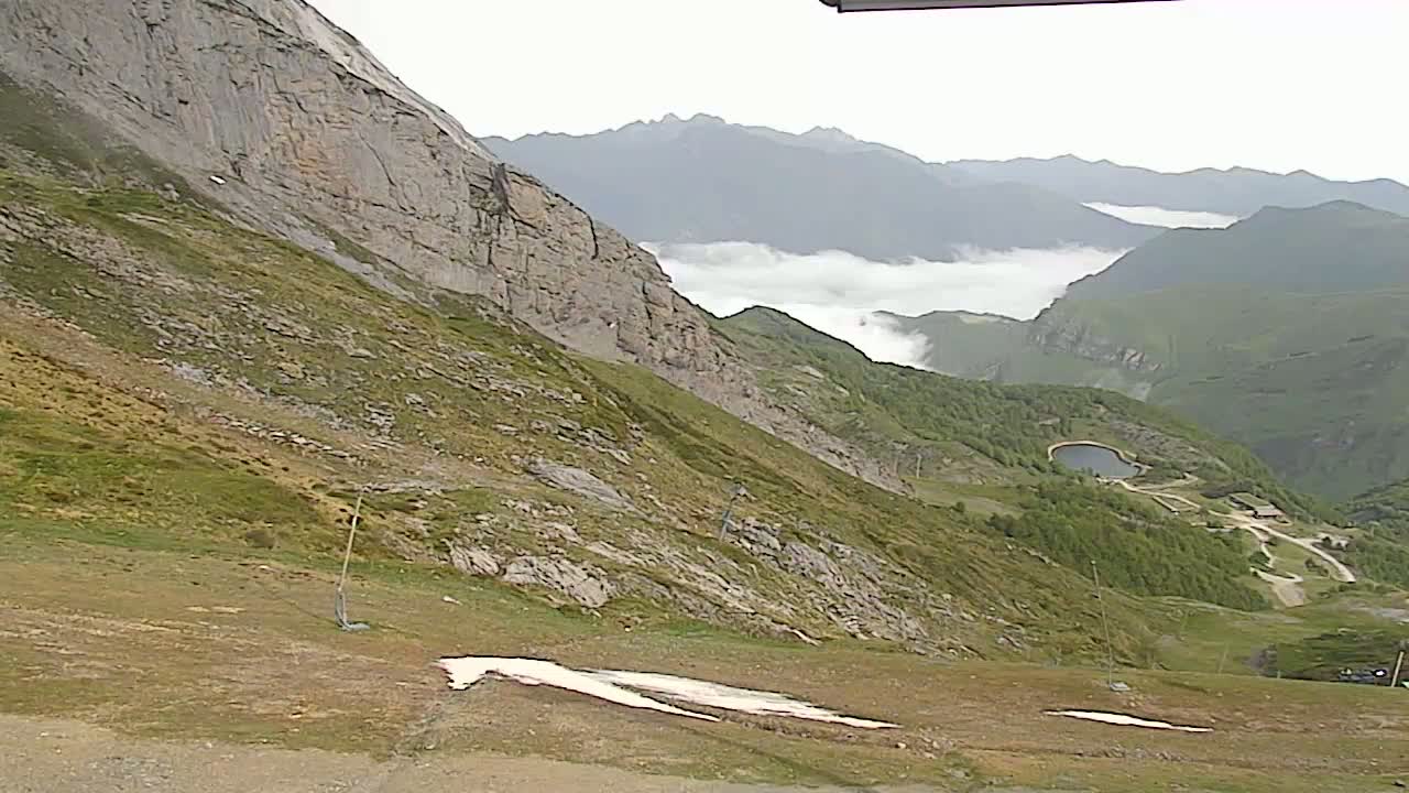 A mountainside with patchy grass and small remnants of snow overlooks a valley partially shrouded in fog and containing a small lake, under an overcast sky.