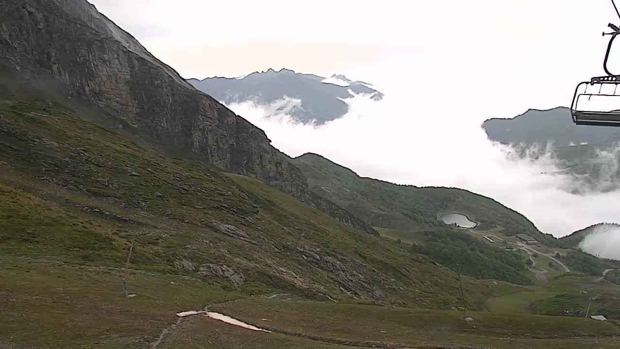 A chairlift ascends a mountainous landscape shrouded in low-lying clouds, overlooking a valley with a small body of water.