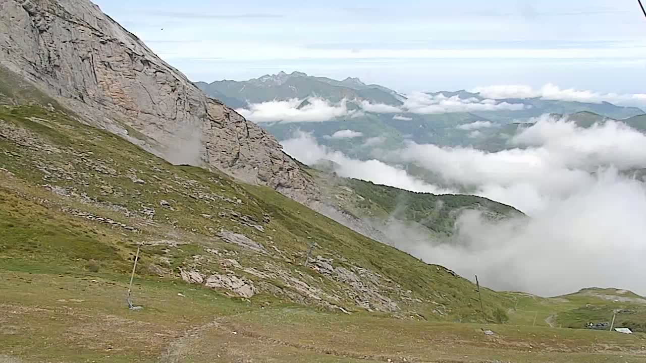 A grassy mountain slope descends towards a valley filled with low-lying clouds and distant mountains under a mostly cloudy sky.