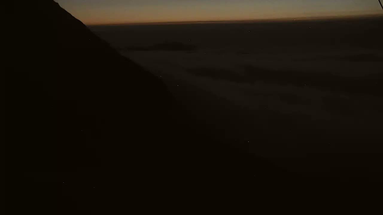 A dark mountainside is silhouetted against a twilight sky above a layer of clouds.