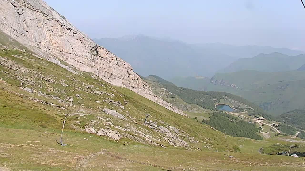 A hazy mountain slope with sparse vegetation descends toward a valley containing a small lake and a few buildings.