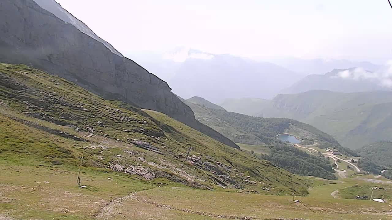 A hazy mountainside scene reveals a steep grassy slope leading down to a valley with a small lake and some structures, all under a mostly clear sky.