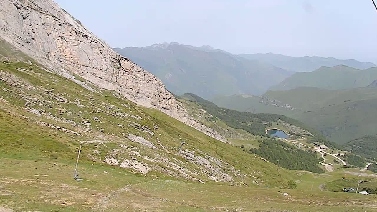 A grassy mountain slope descends toward a valley with a small lake and distant mountains under hazy, sunny conditions.