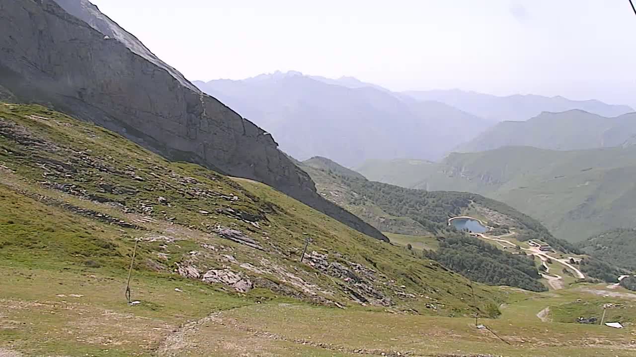 A hazy mountain vista reveals a steep, grassy slope descending towards a valley with a small lake and scattered buildings, under a bright, clear sky.