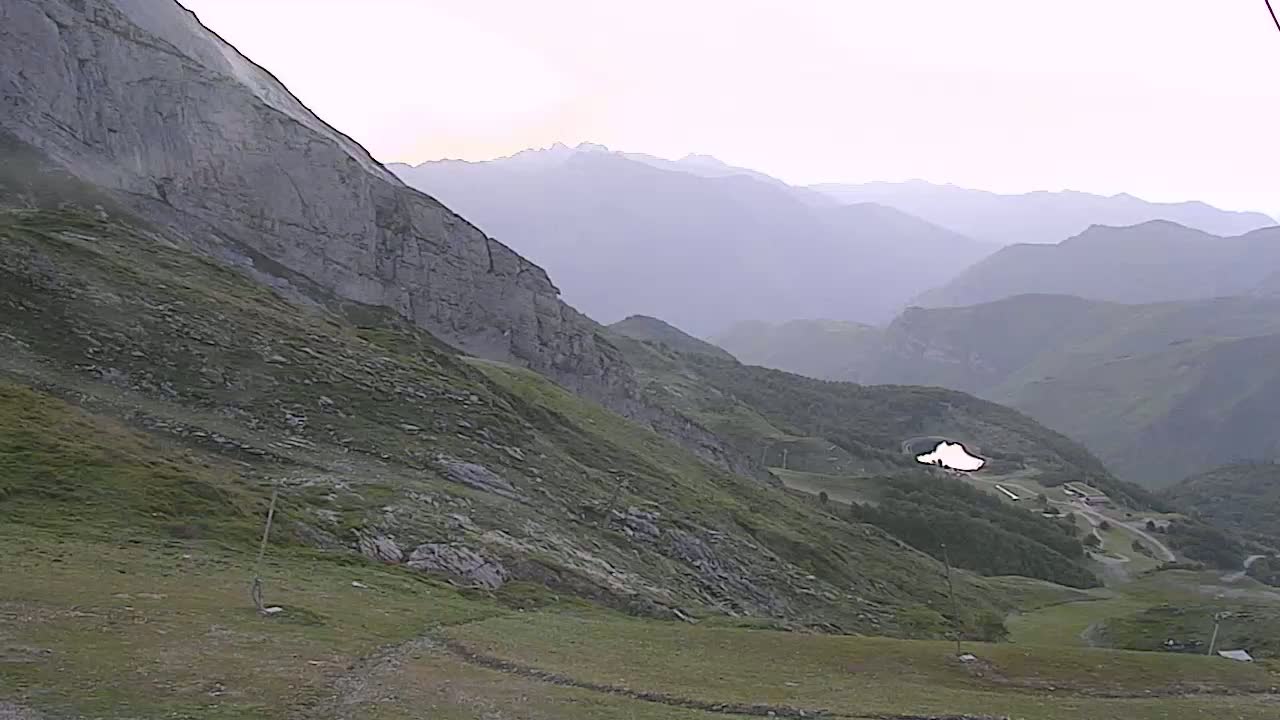 A hazy mountain landscape shows a steep, rocky hillside descending to a valley with a small lake and distant mountains under an overcast sky.
