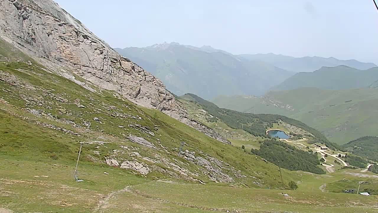 A grassy mountain slope descends to a valley with a small lake and winding roads, under a hazy sky.