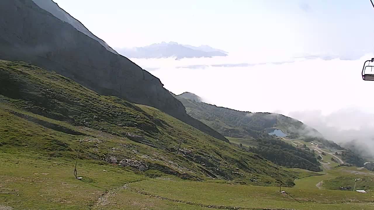 A grassy mountain slope descends towards a valley partially obscured by fog, with a chairlift visible in the upper right corner.