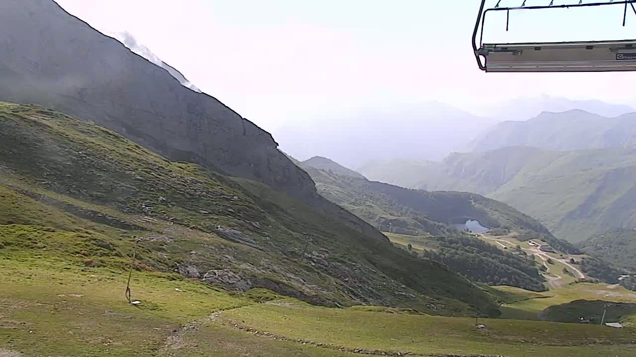 A chairlift extends over a grassy mountainside overlooking a hazy valley with distant mountains under a bright, clear sky.