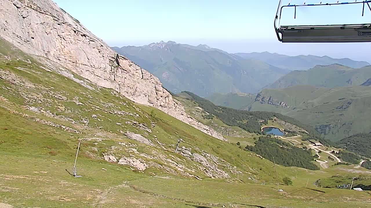 A chairlift ascends a grassy mountain slope overlooking a valley with a small lake and distant hazy mountains under a clear sky.