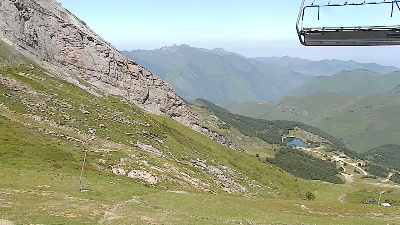 A chairlift ascends a grassy mountain slope overlooking a valley with a small lake and distant mountains under a clear sky.