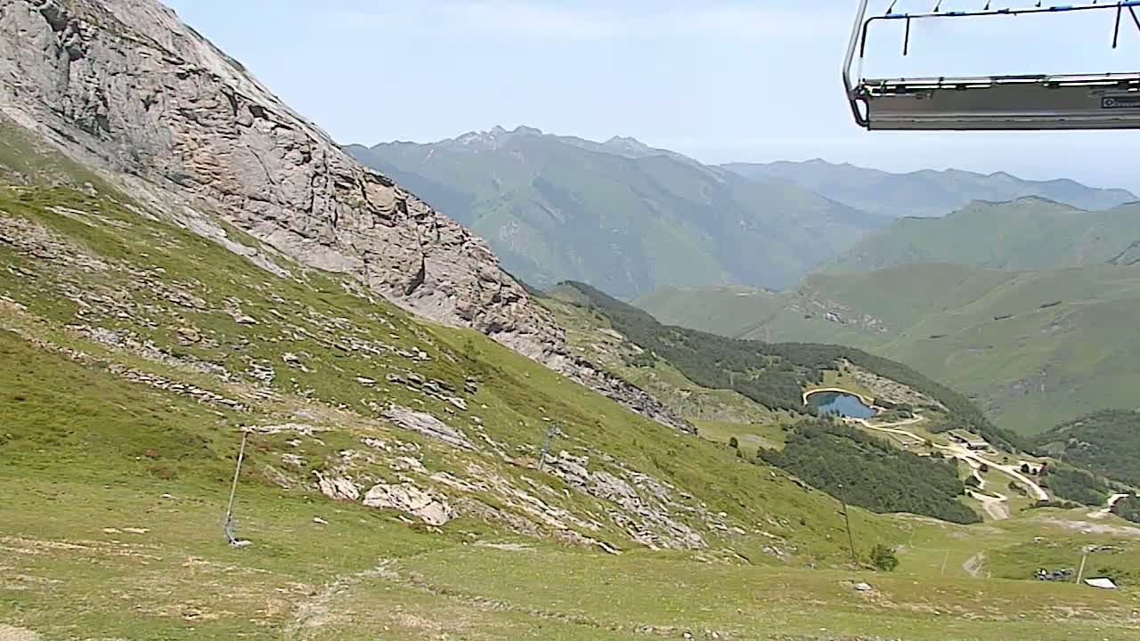 A chairlift ascends a grassy mountain slope overlooking a valley with a small lake and distant mountains under a clear sky.
