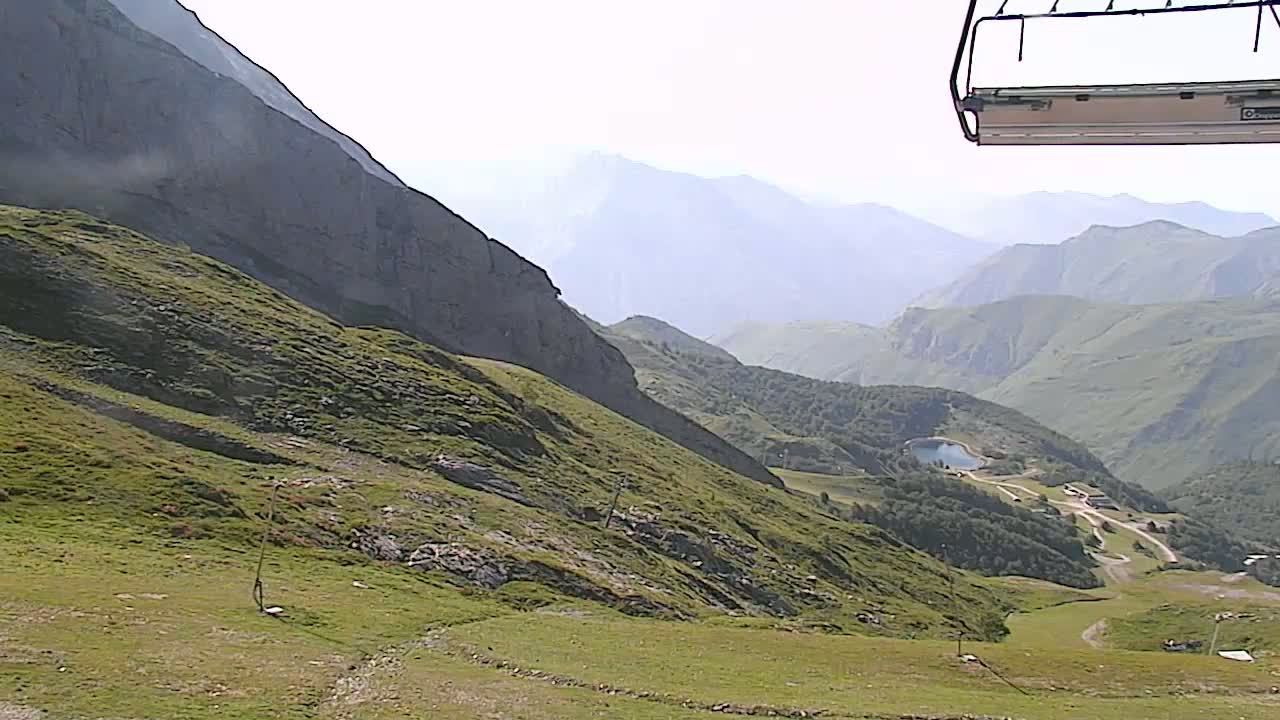 A chairlift ascends a grassy mountain slope overlooking a valley with distant hazy mountains under a bright, clear sky.