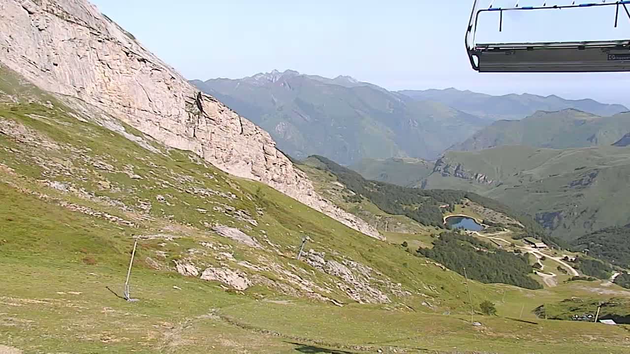 A chairlift ascends a grassy mountain slope overlooking a valley with a small lake and distant mountains under sunny skies.