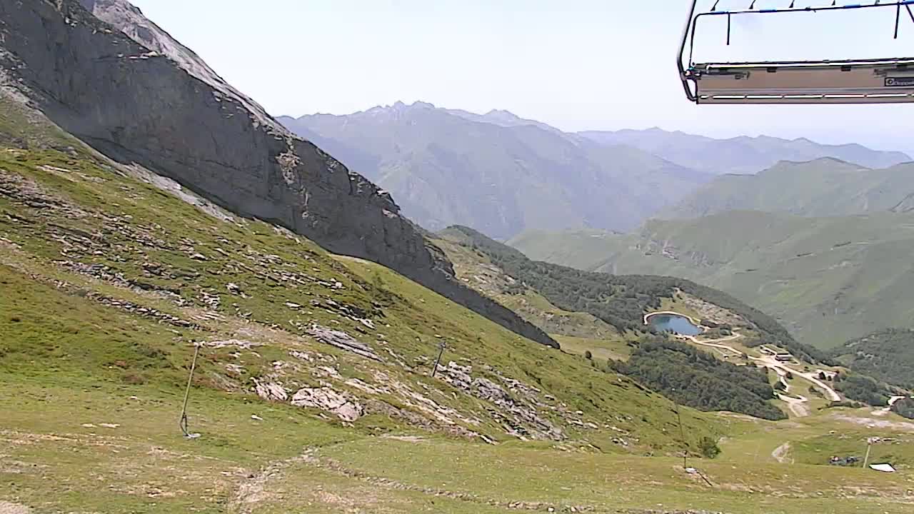 A mountainside with sparse vegetation and rocky outcroppings slopes down to a valley with a small lake and a few buildings under a clear, sunny sky, partially obscured by a chairlift.