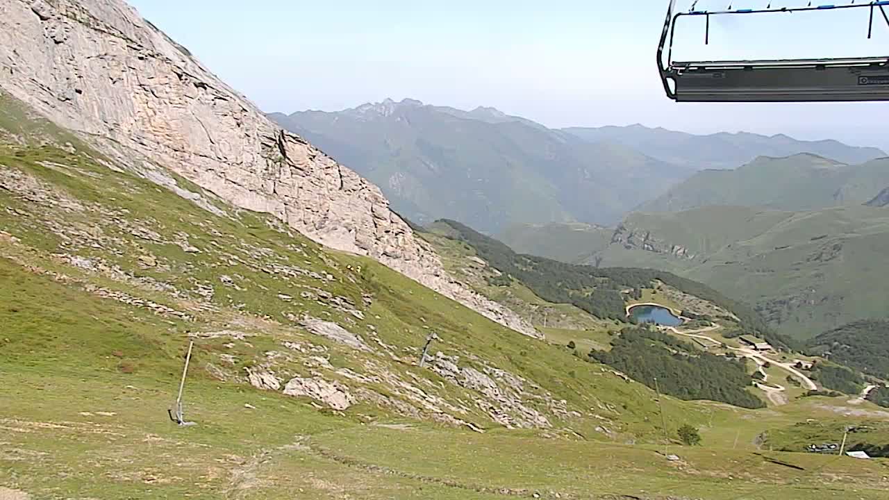 A chairlift ascends a grassy mountain slope overlooking a valley with a small lake and distant mountain ranges under a clear sky.