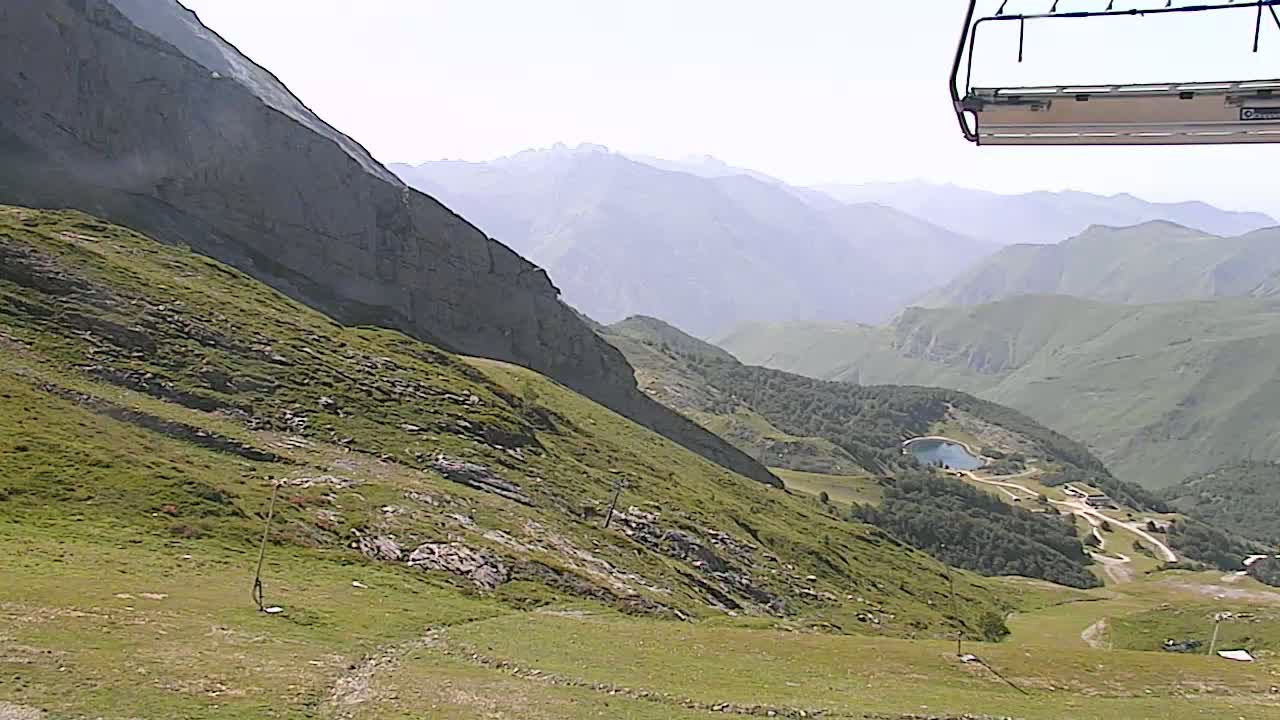 A chairlift hangs over a grassy mountain slope overlooking a valley with a small lake and distant mountains under a bright, hazy sky.