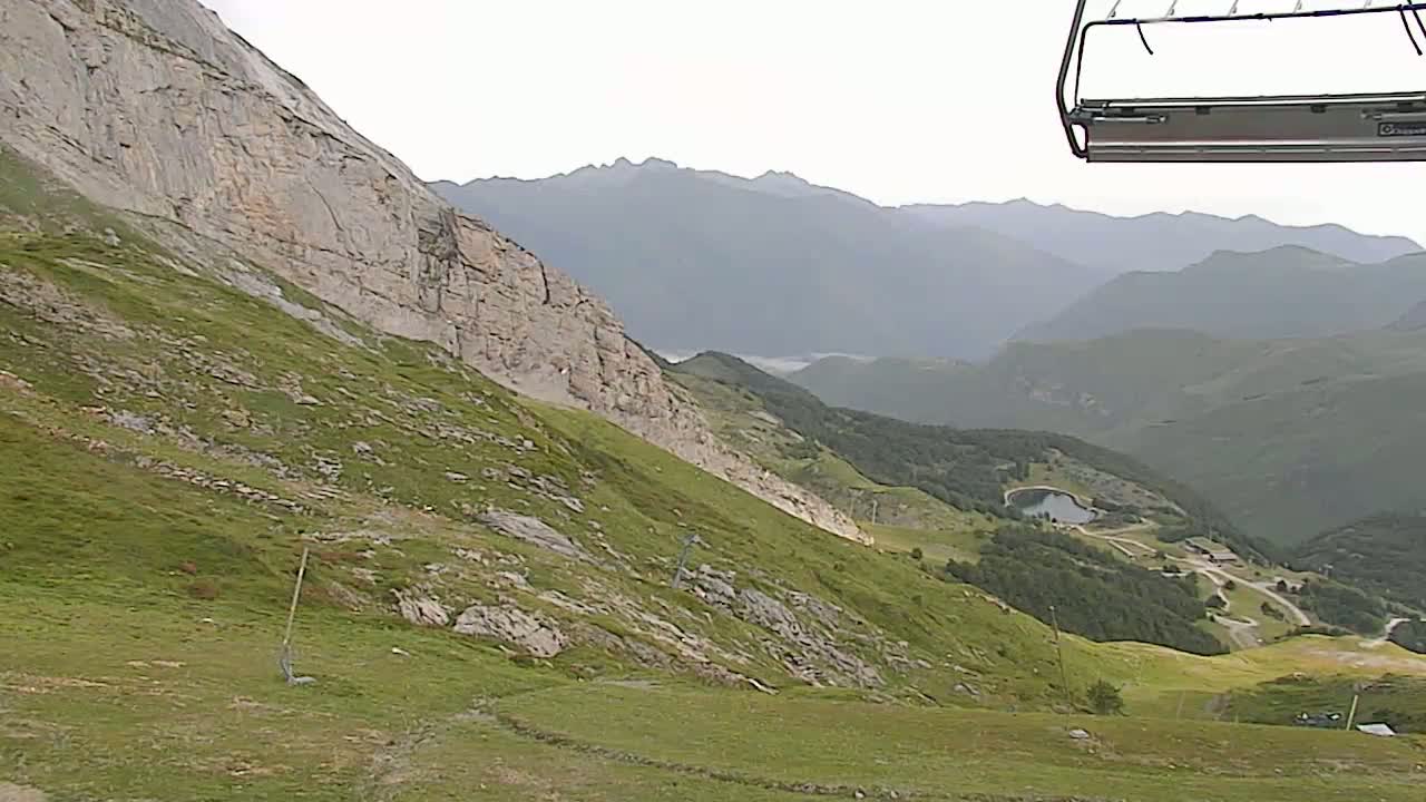 A chairlift ascends a grassy mountain slope overlooking a valley with a small lake under an overcast sky.